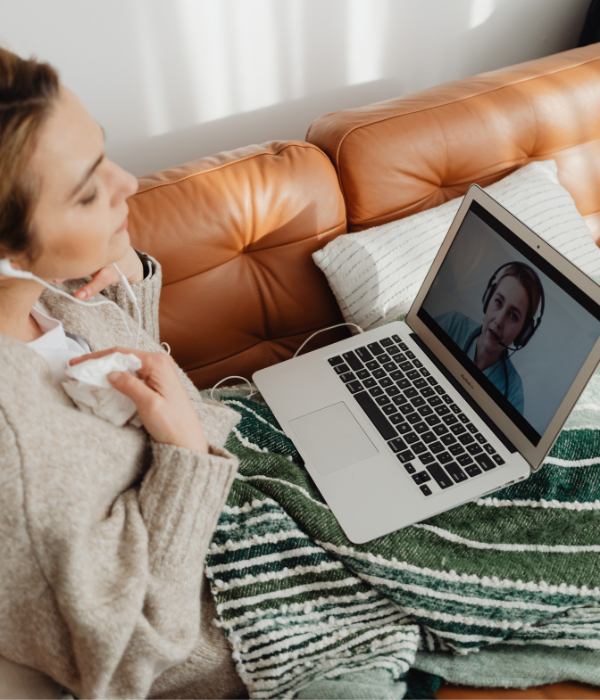 A woman sits on a couch with a laptop in a video meeting with an online therapist.