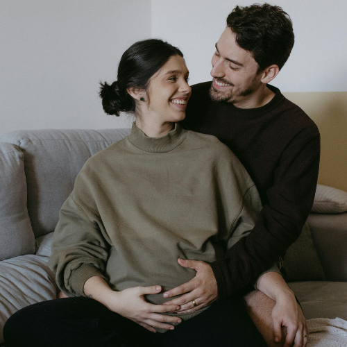 A young man and woman couple, who are expecting a baby, sit on a couch. They are smiling because online couples therapy has helped them grow closer as they welcome every new chapter together.