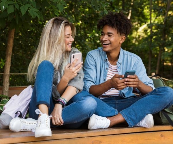 A white teenage girl and a black teenage boy sit close and laugh with their phones in their hands depicting how young love can be fun but challenging. Teen counseling can help teens process their confusing emotions.